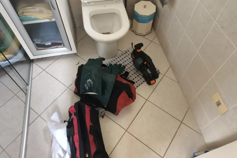 Banheiro com bolsas de ferramentas e luvas no piso ao lado do vaso sanitário