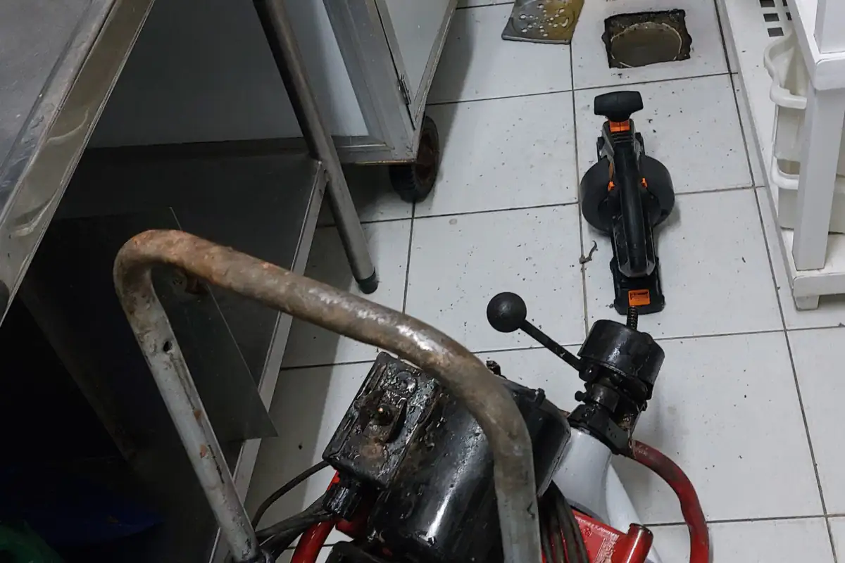Cozinha industrial com bancada de inox, ralo de piso aberto e máquina de desentupimento sobre o piso de azulejo