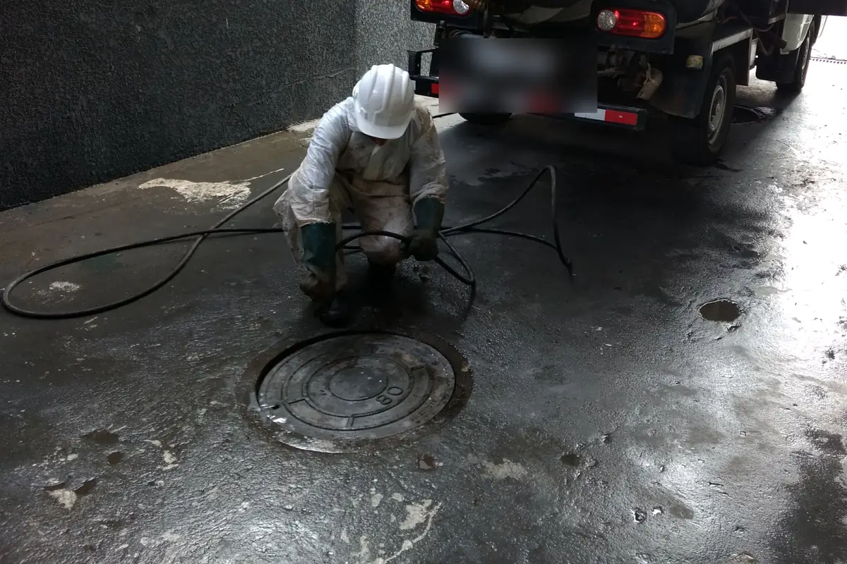 Técnico de capacete branco e macacão agachado ao lado de uma tampa de ferro fundido fechada, com mangueira preta no chão