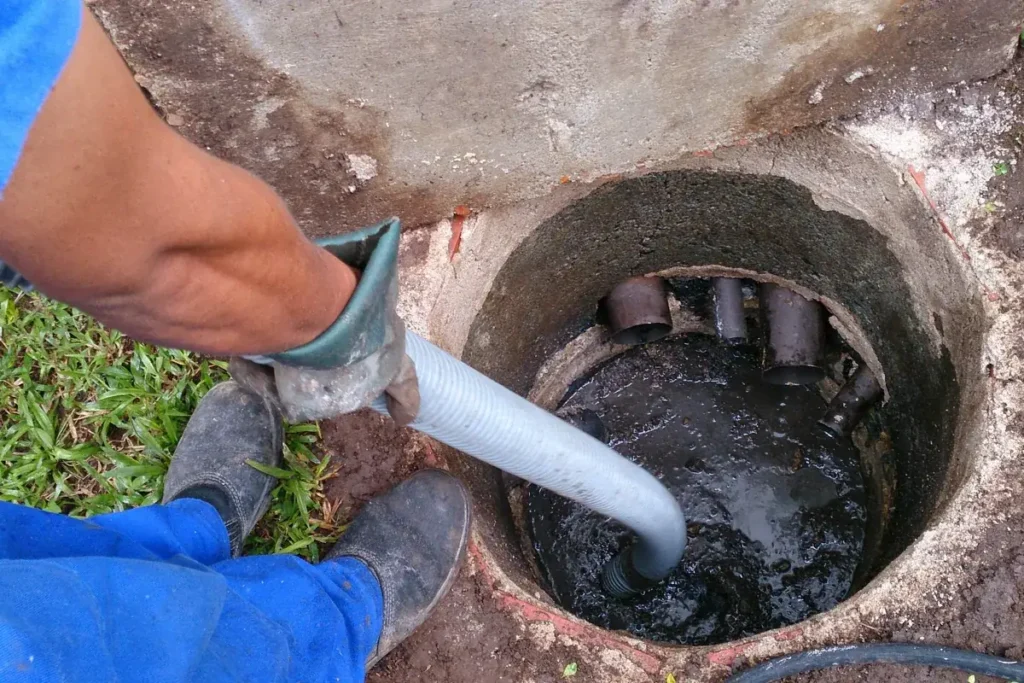 Técnico de macacão azul e luvas retirando o conteúdo de uma fossa de concreto aberta com mangueira de sucção