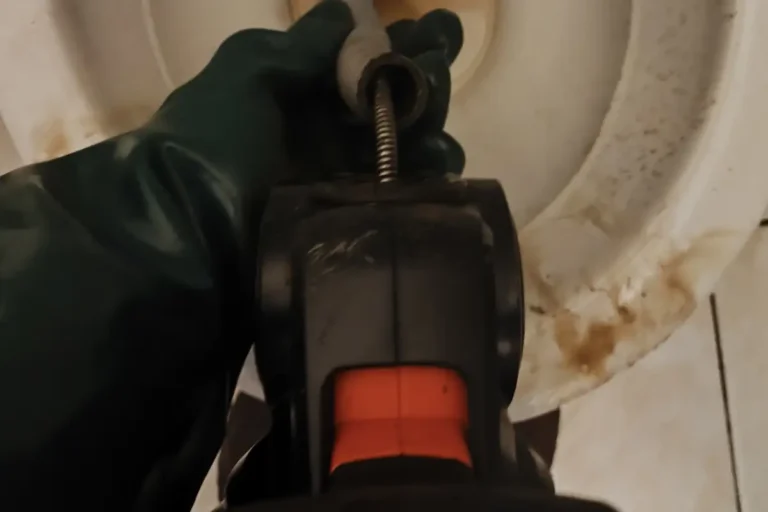 Mão com luva verde segurando a máquina de desentupimento manual dentro da bacia do vaso
